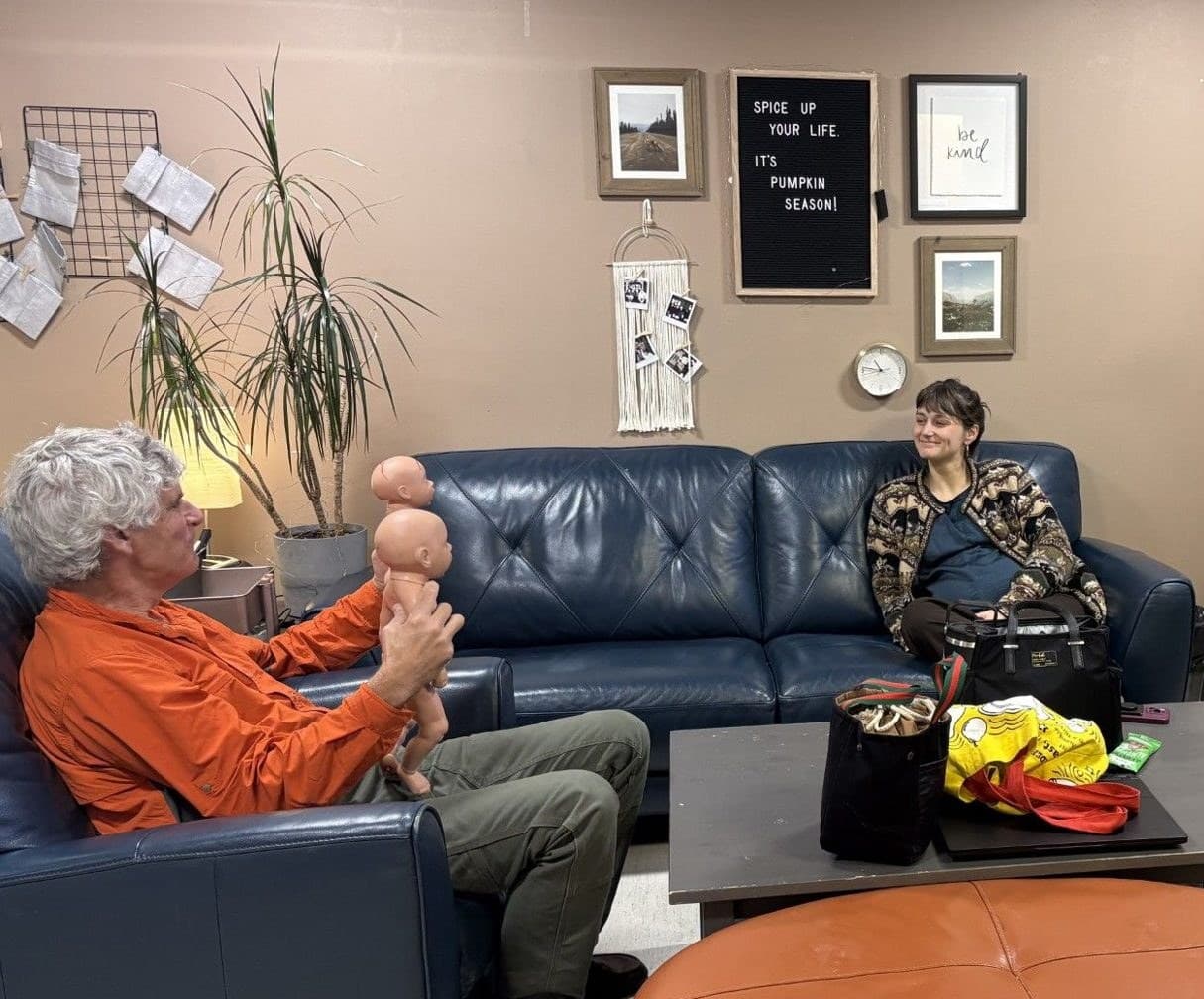 A faculty member in an orange shirt holds up two newborn simulation dolls to teach, while a resident on the couch listens in the resident lounge