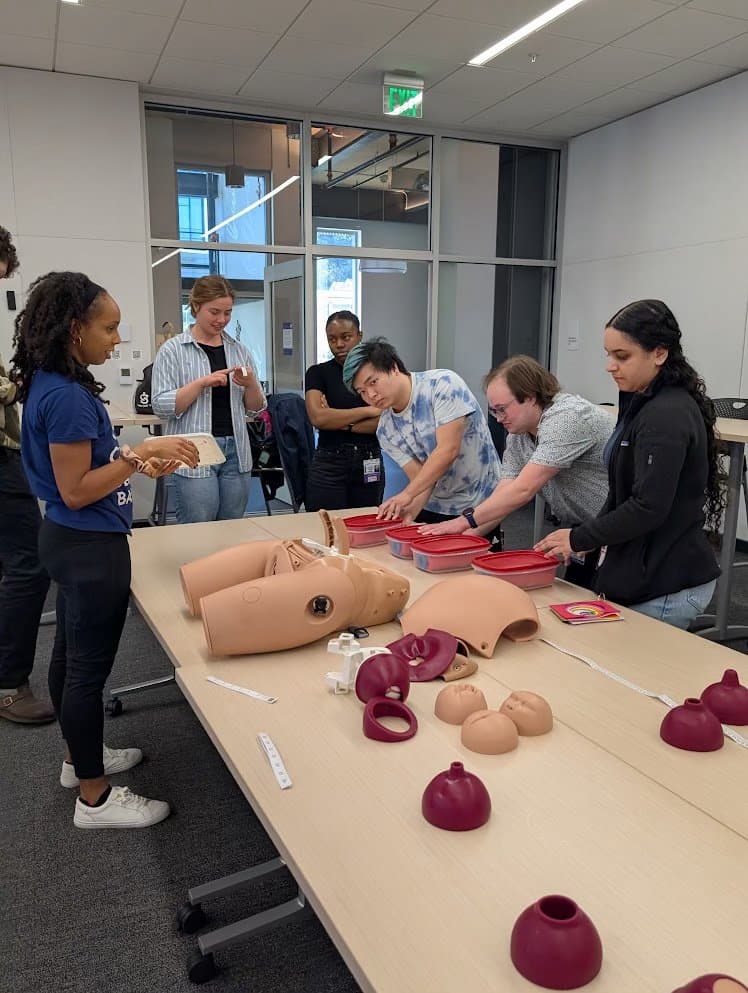 Residents examining pelvic and birthing simulation models laid out on a long table while a facilitator explains