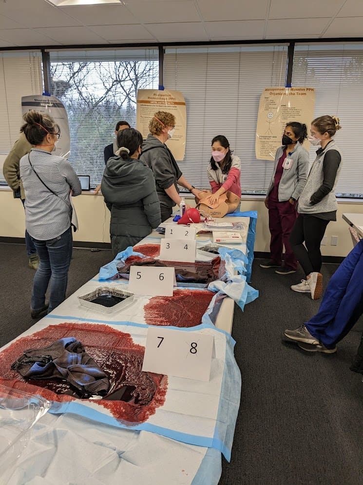 Residents and faculty at numbered stations with simulated blood loss on absorbent pads, beneath posters on postpartum hemorrhage and shoulder dystocia