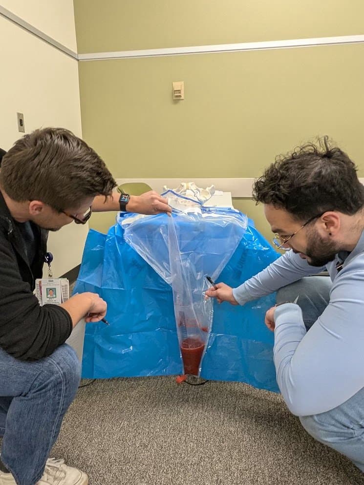 Two residents crouching beside an under-buttocks drape, reading the measured blood loss in its graduated collection pouch