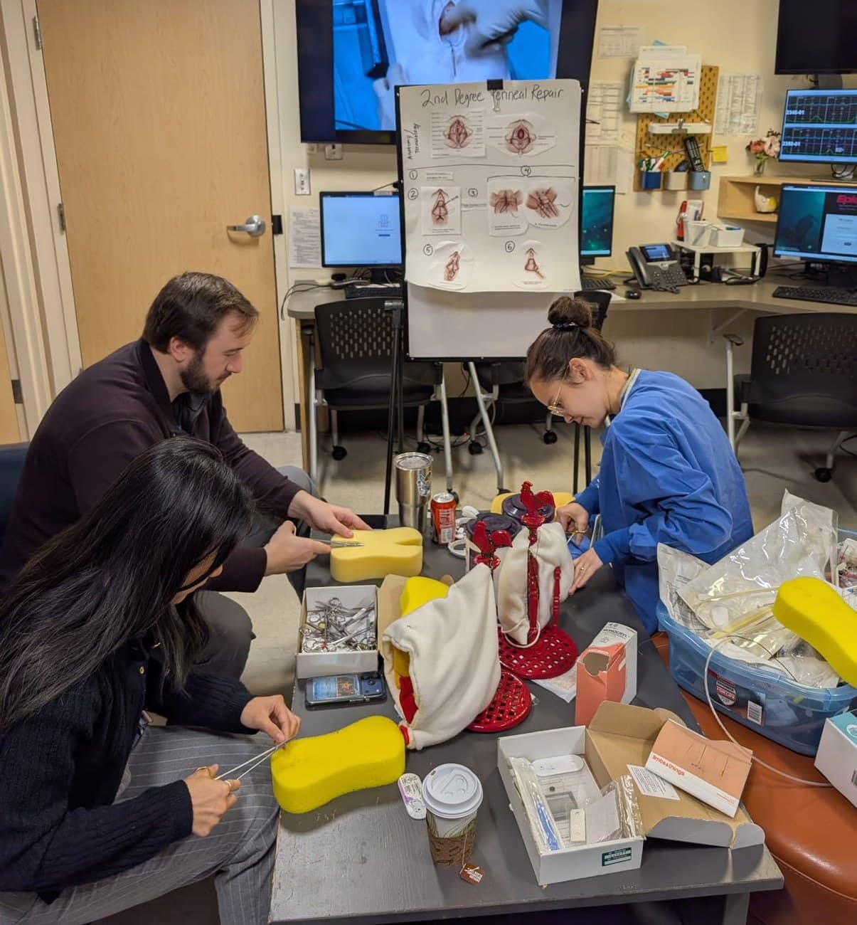 Three residents practicing laceration repair on sponge and fabric perineal models at a table, with a second-degree perineal repair flip chart behind them