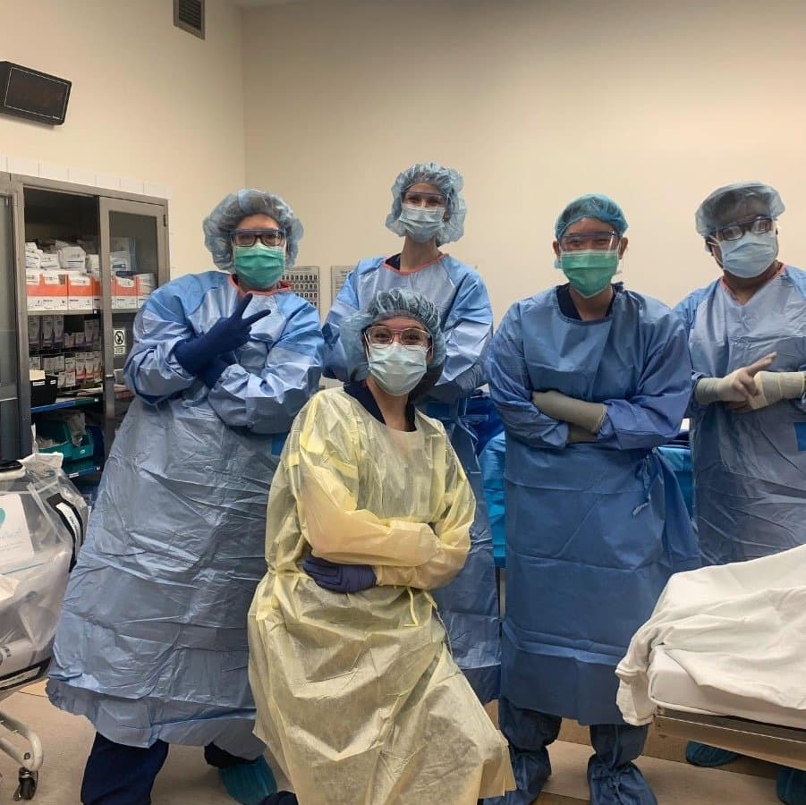 Five residents in surgical gowns, caps, masks and eye protection posing together in an operating room