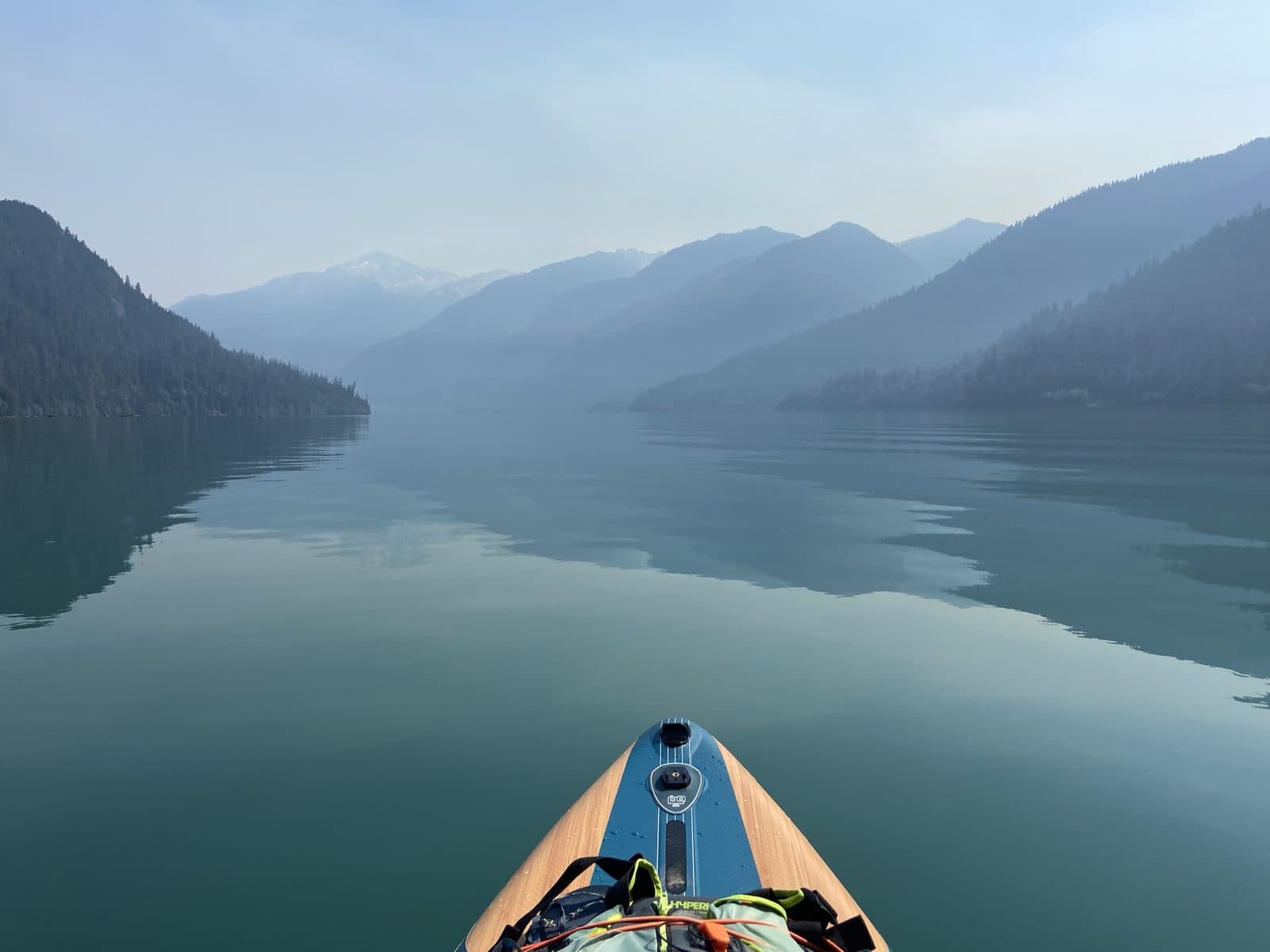 The nose of a paddleboard on a still, glassy lake between forested mountains