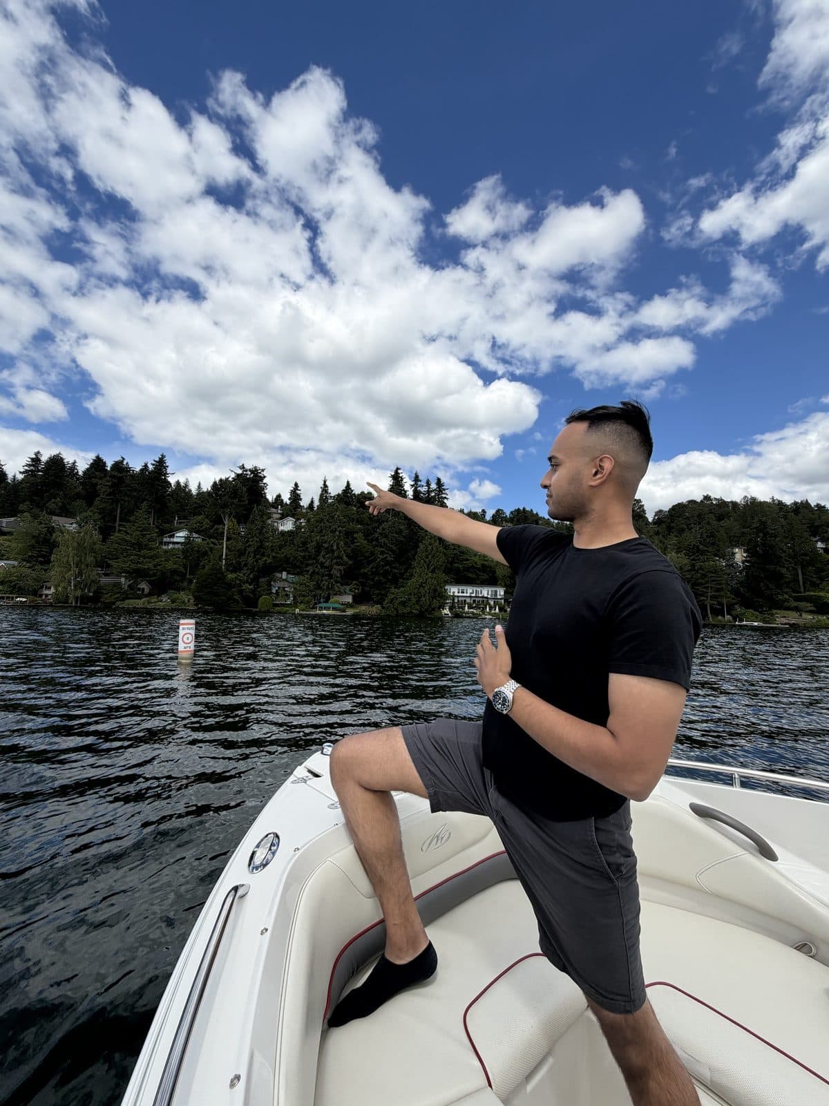 A resident standing at the bow of a boat pointing toward a wooded shoreline under a bright, cloudy sky