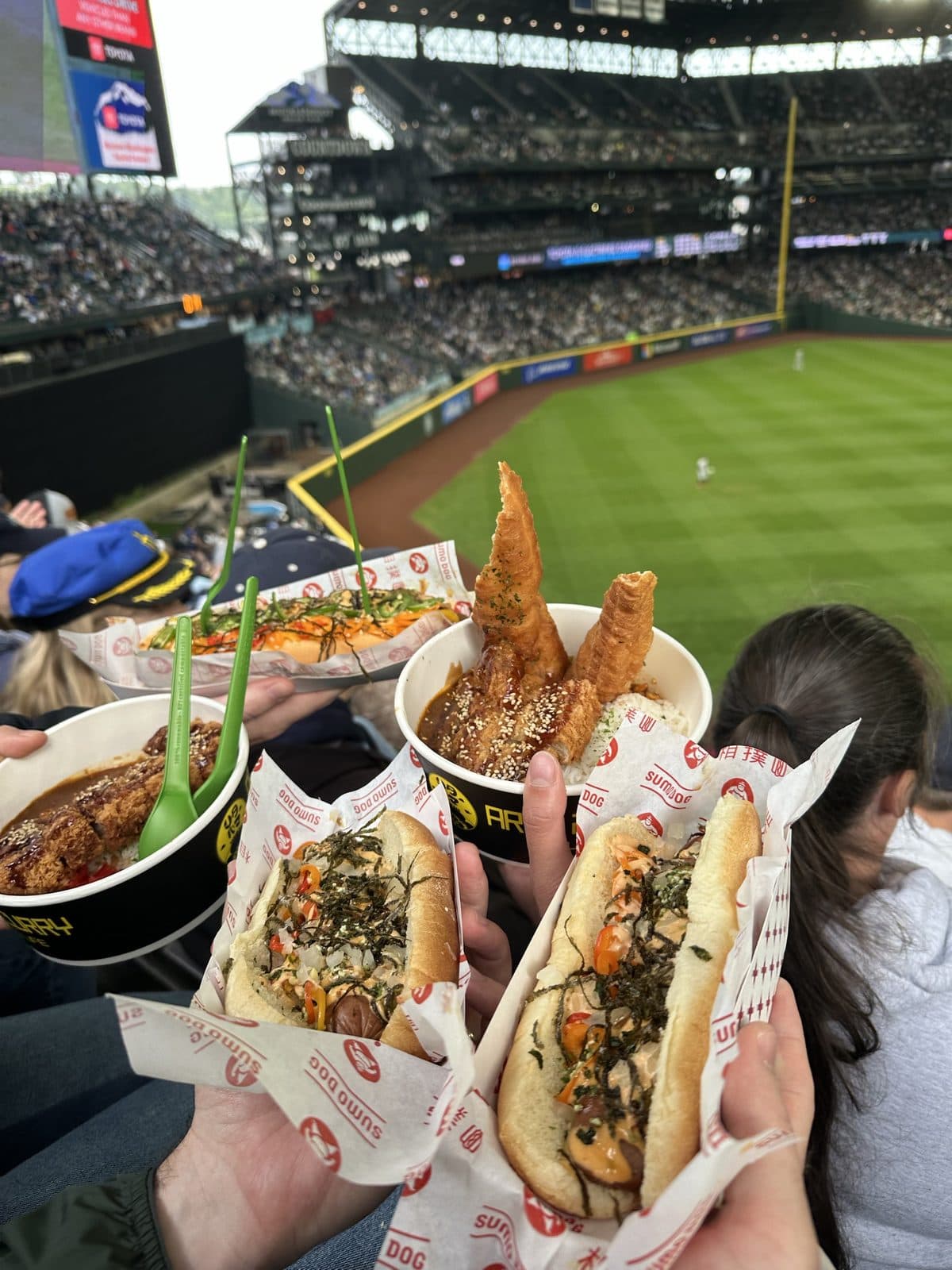 Hot dogs and katsu curry bowls held up in the stands above the outfield at a Mariners game