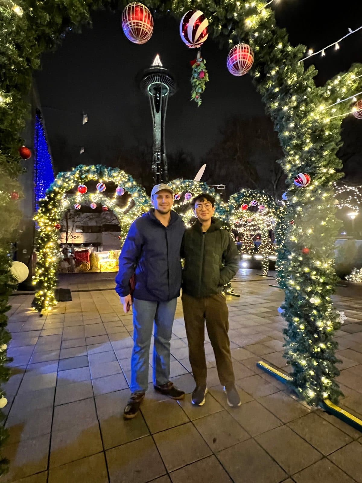 Two residents under arches of holiday lights and ornaments at Seattle Center, with the Space Needle behind them