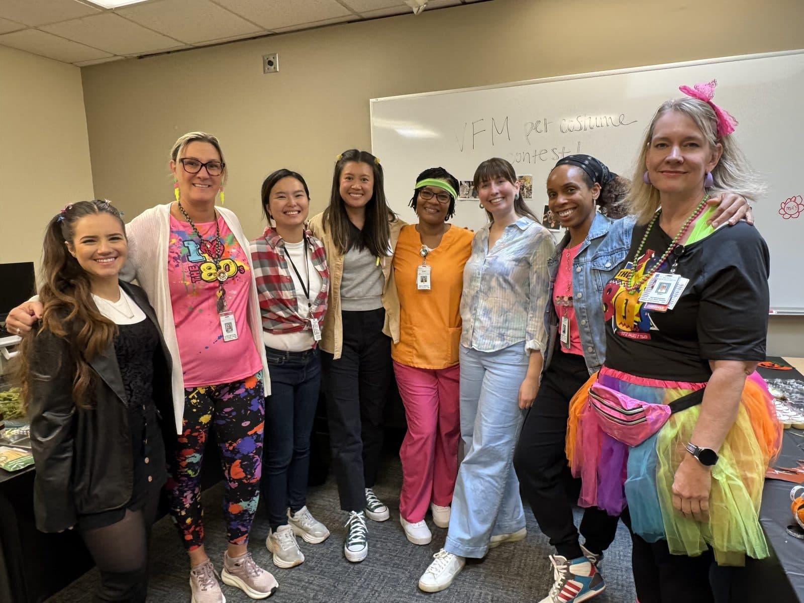 Faculty and staff in neon 1980s outfits, tutus and leg warmers in front of a whiteboard reading VFM pet costume contest