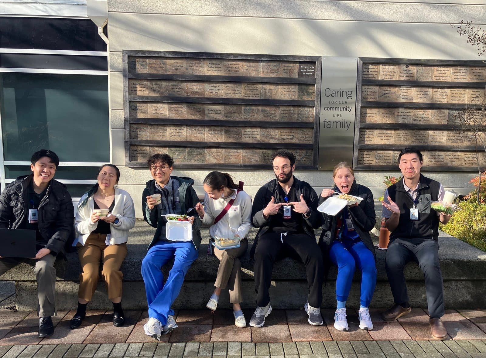 Seven residents making silly faces while eating lunch on a bench beside a wall reading Caring for our community like family