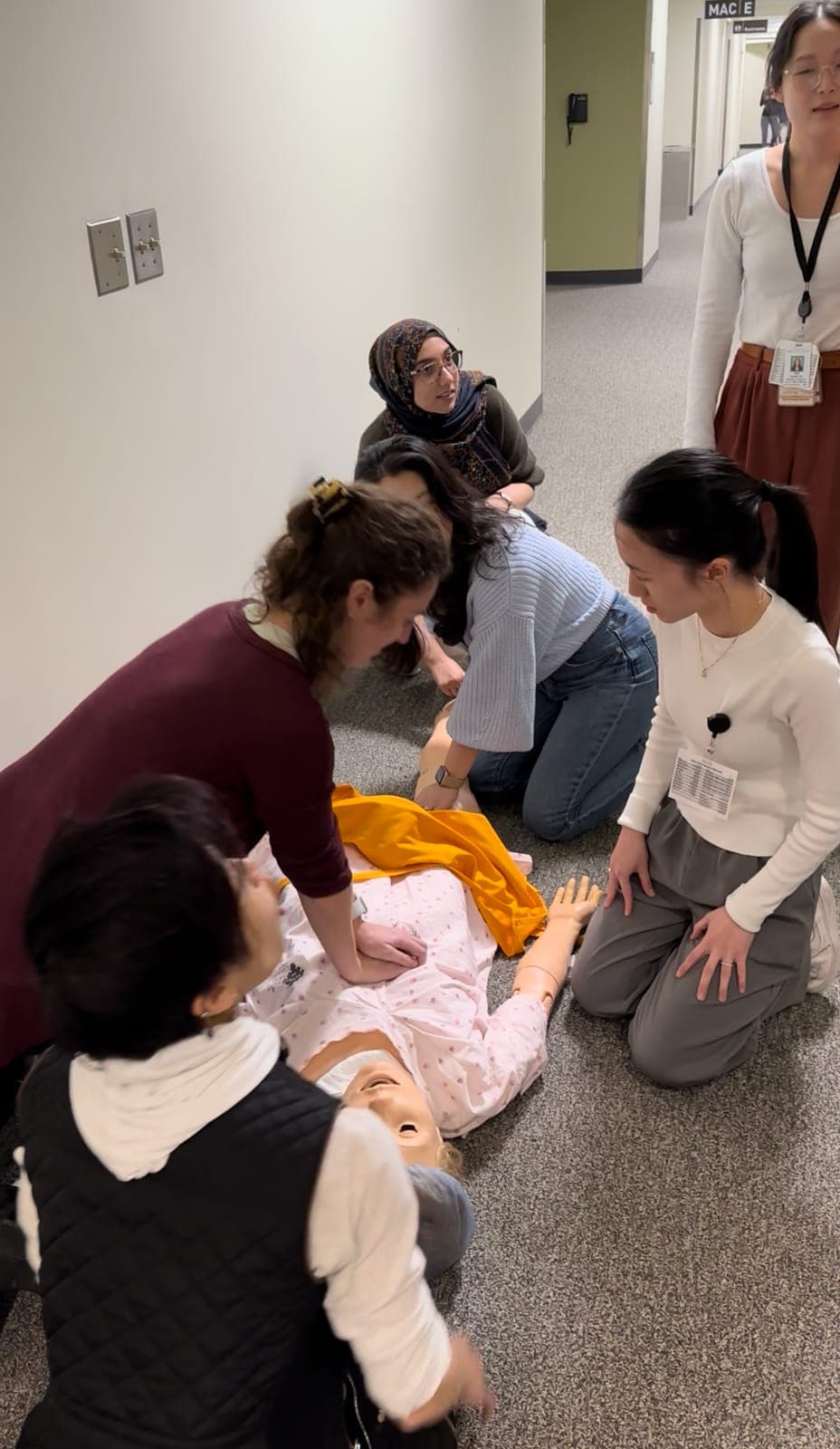 Residents kneeling around a simulation mannequin on a hallway floor while one performs chest compressions