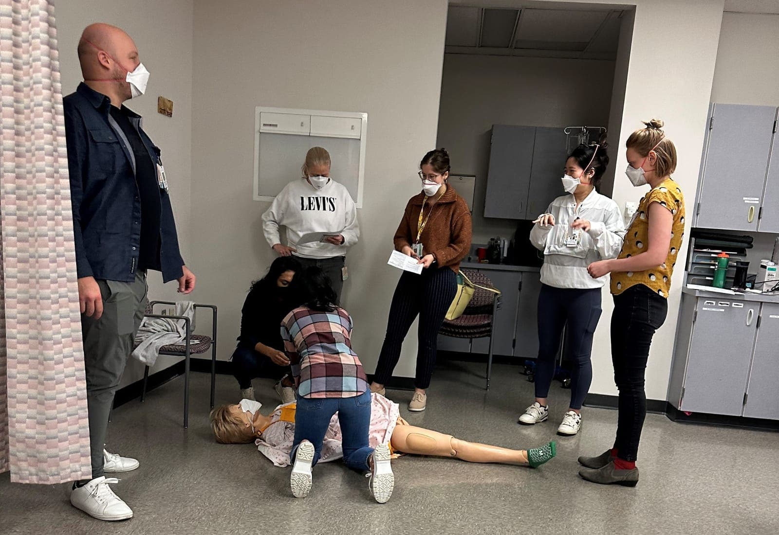 A resident performing chest compressions on a mannequin on the floor of a clinic room while faculty and residents watch with clipboards
