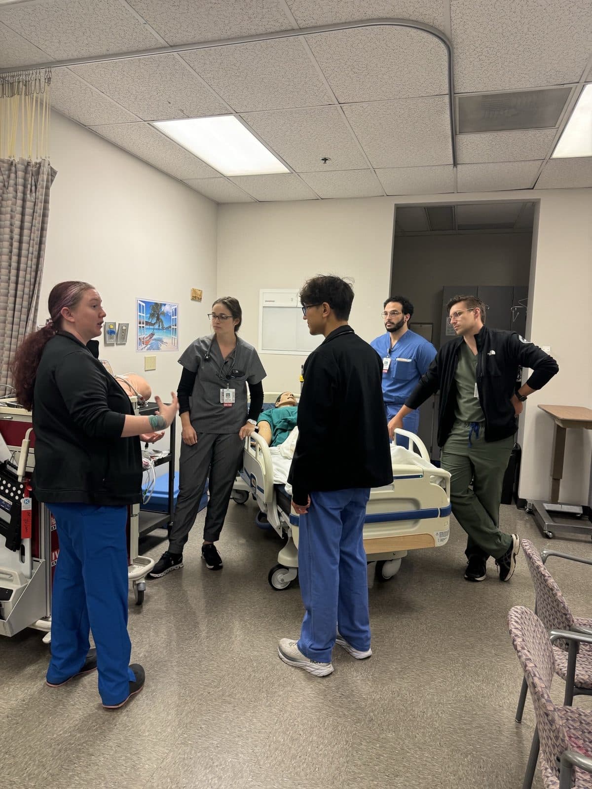 A facilitator debriefing residents in scrubs beside a simulation mannequin in a hospital bed in the simulation lab