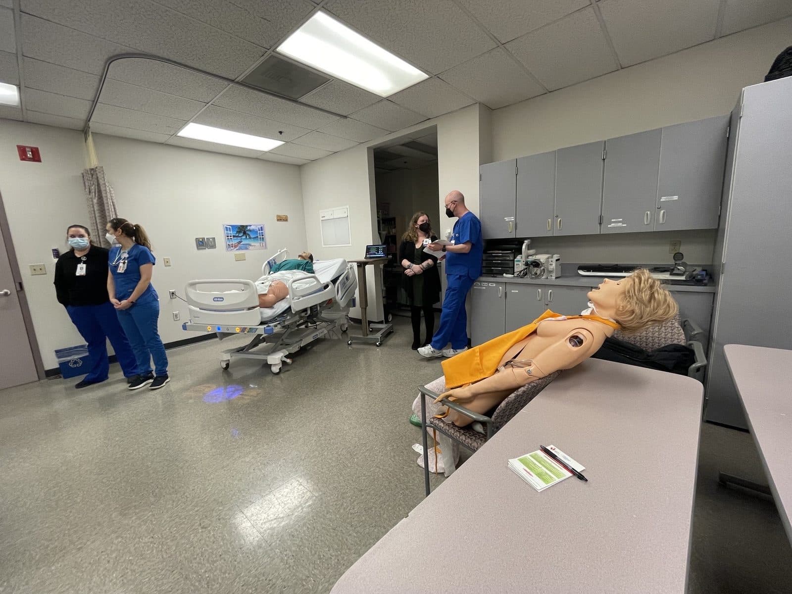 Residents and facilitators in the simulation lab with a mannequin in a hospital bed and another seated in a chair