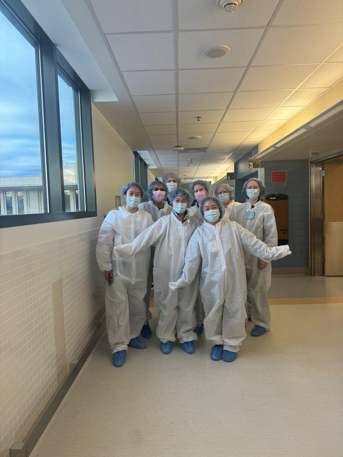 Residents in white protective coveralls, masks, and hair and shoe covers posing in an operating room hallway