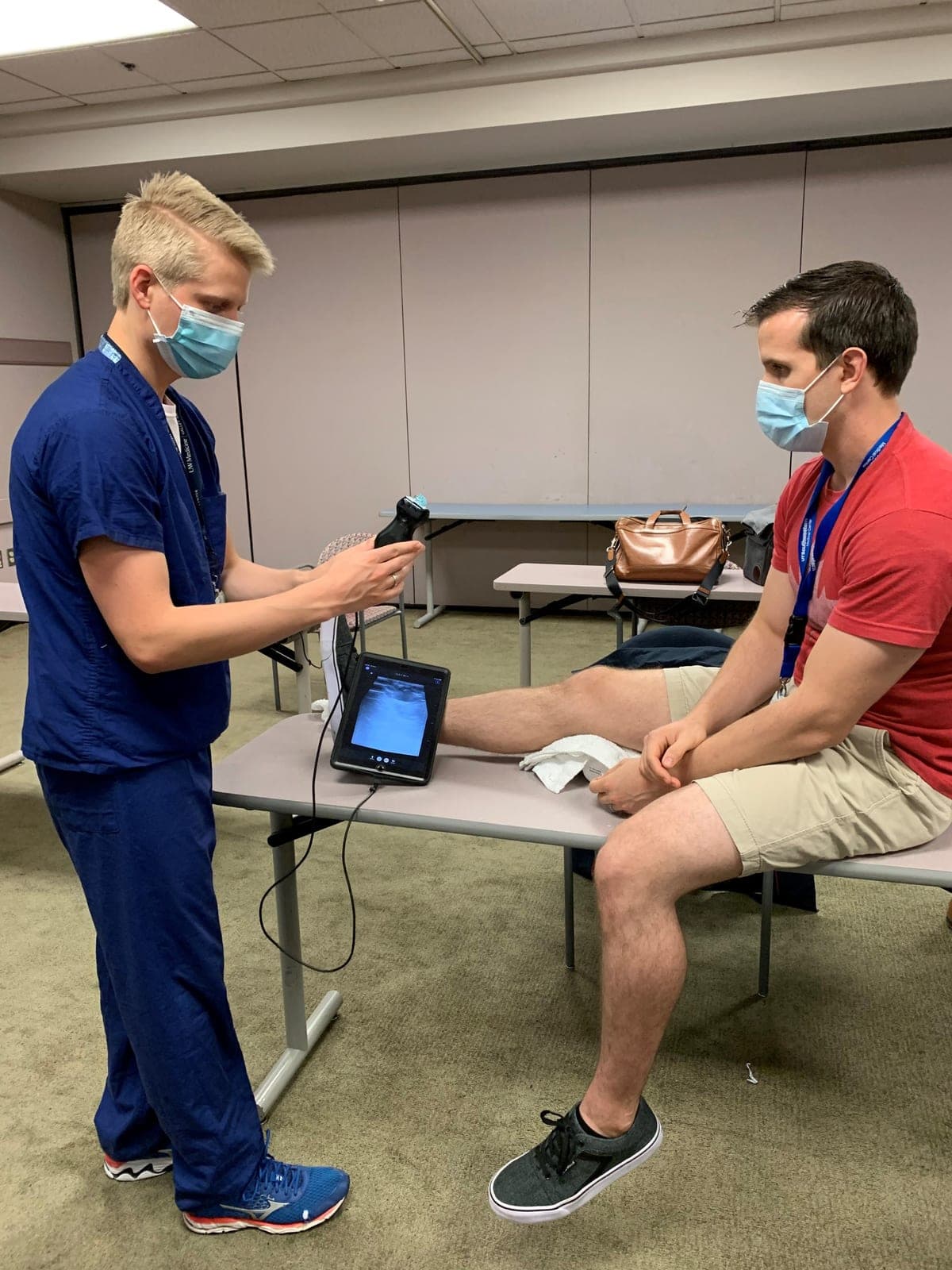 A resident scanning a volunteer's knee with a handheld ultrasound probe, the image on a tablet beside them
