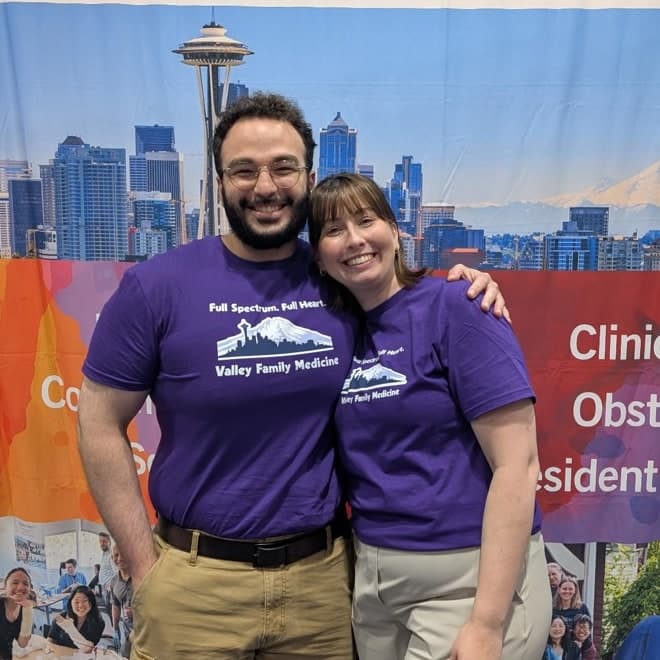 Hamzeh Salti and Kelly Herbig in purple Valley Family Medicine shirts, standing in front of the program's conference banner with the Seattle skyline