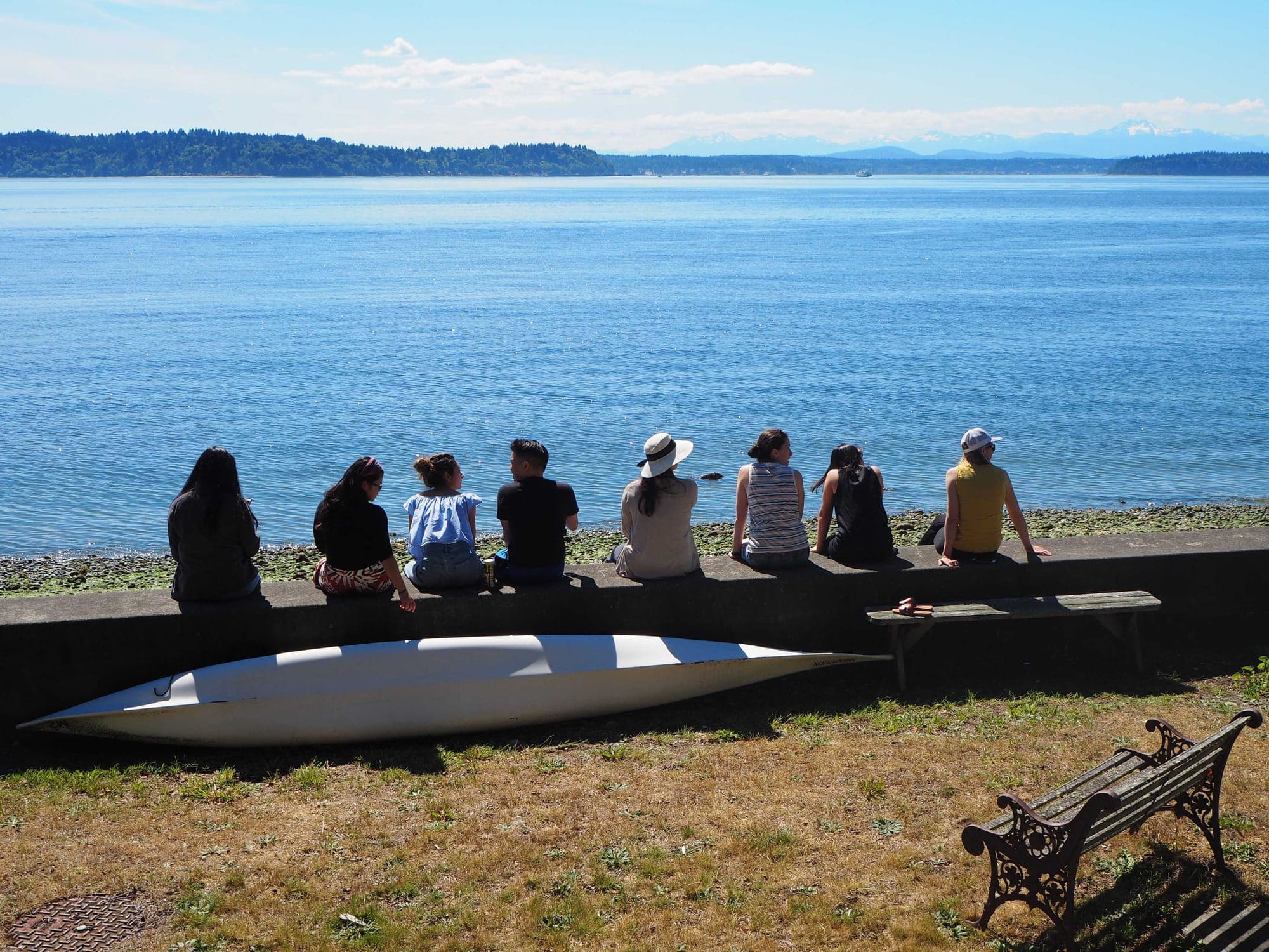 The class of 2024 seen from behind, sitting on a sea wall looking out over Puget Sound