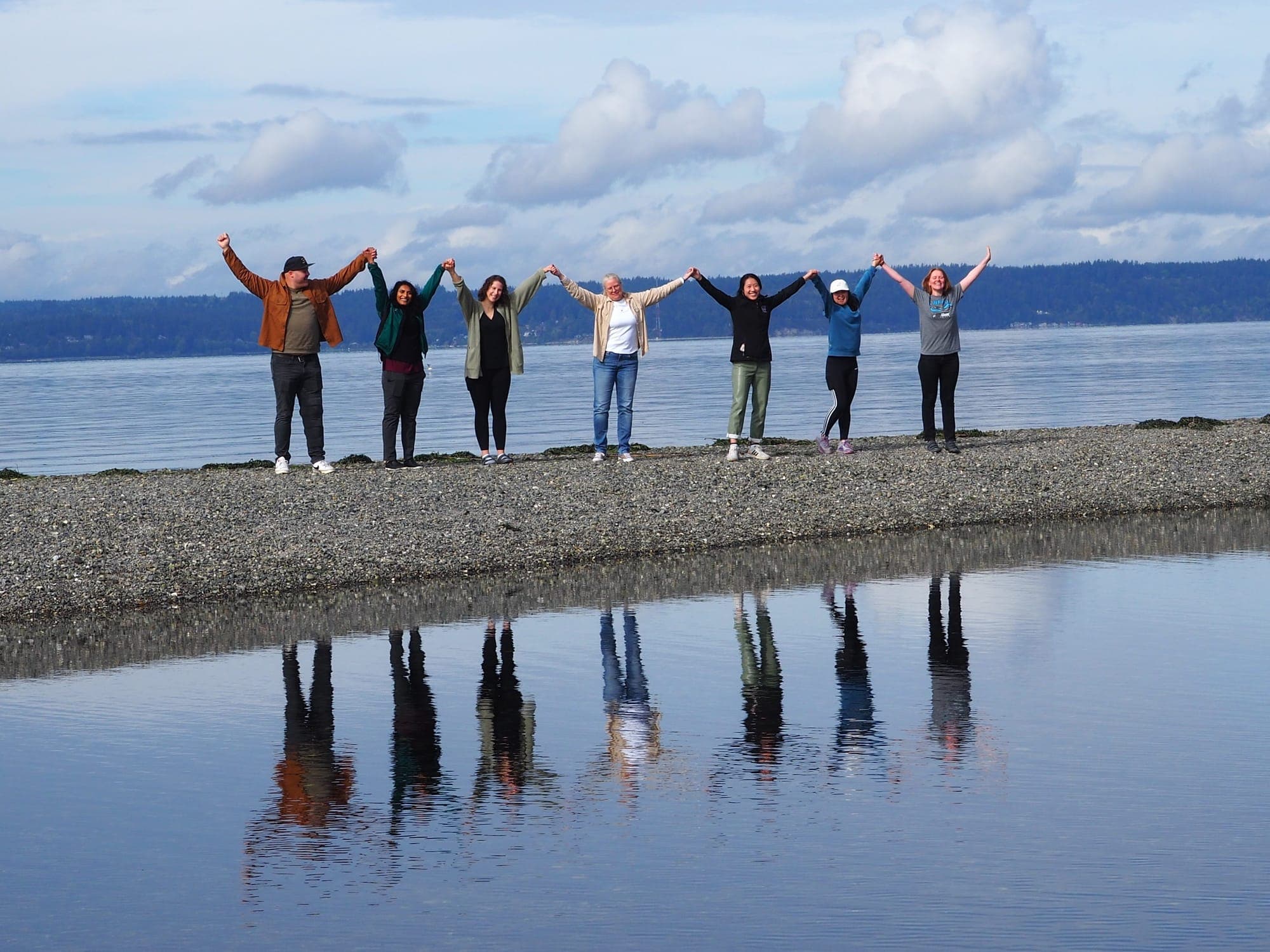 The class of 2025 standing in a line with arms raised on a gravel spit, reflected in the water of Puget Sound